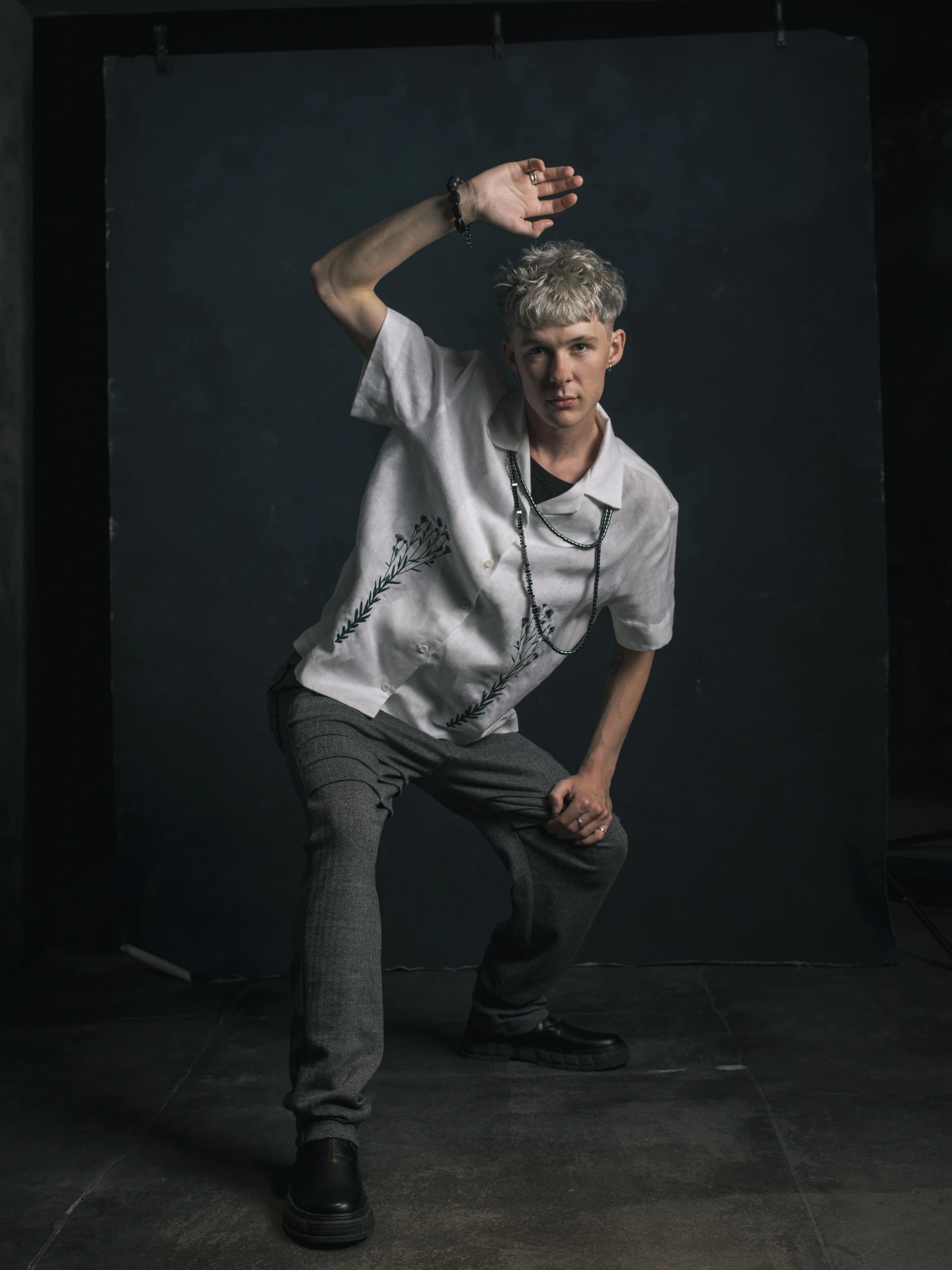 Editorial portrait of a model with platinum blonde hair in a white embroidered linen shirt, posed dynamically against a dark backdrop