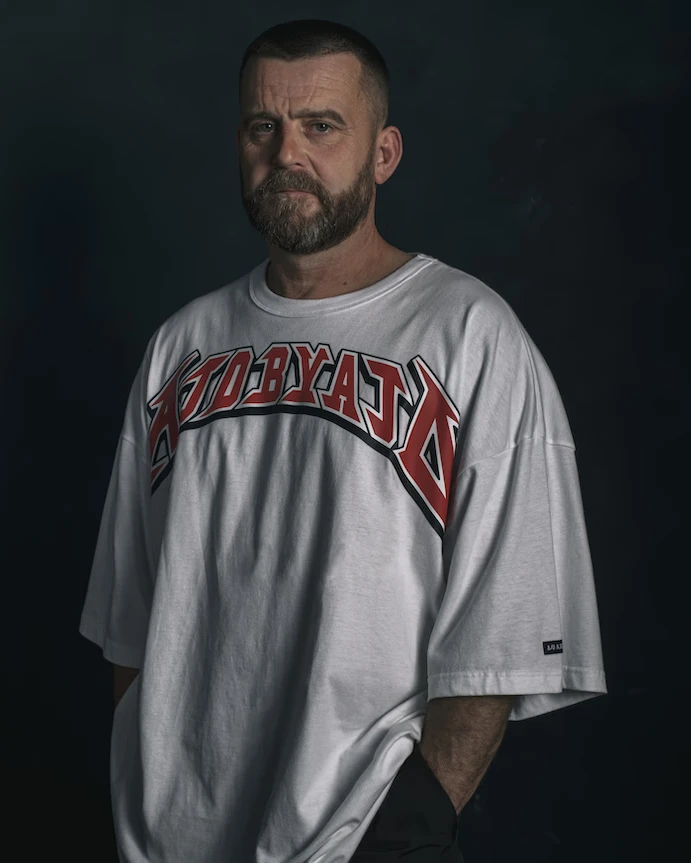 Editorial portrait of a bearded model wearing a graphic oversized tee, photographed against a dark backdrop